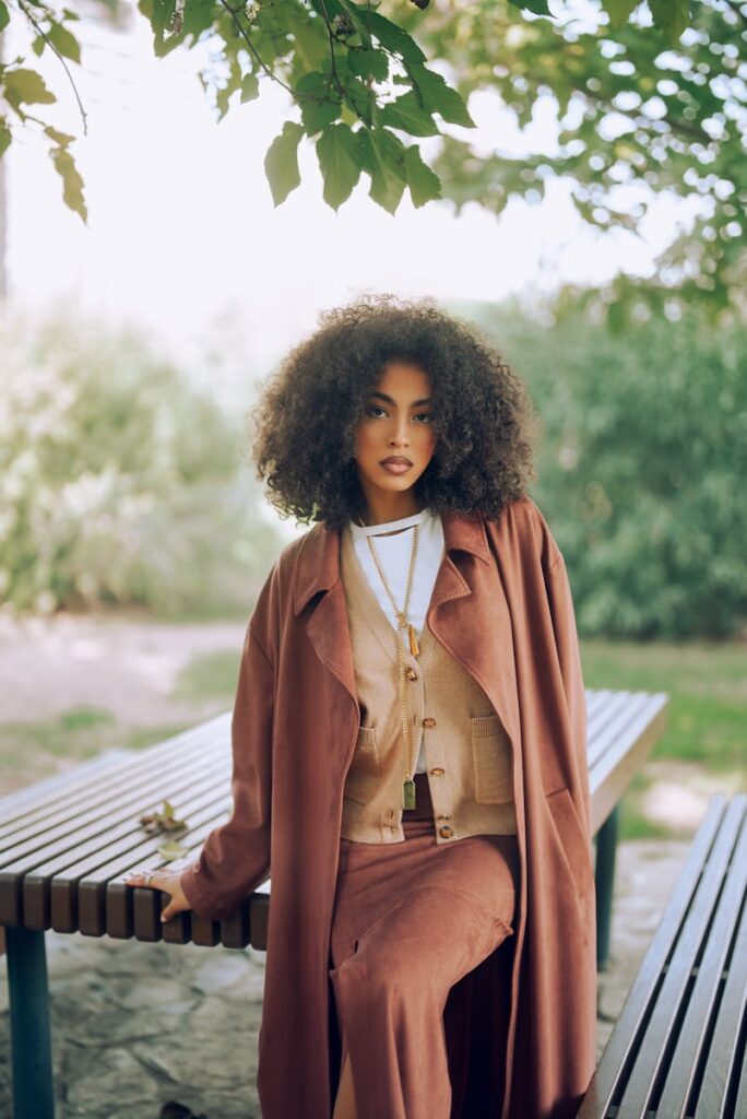 Elegant woman in fall attire sitting on a park bench under lush trees.